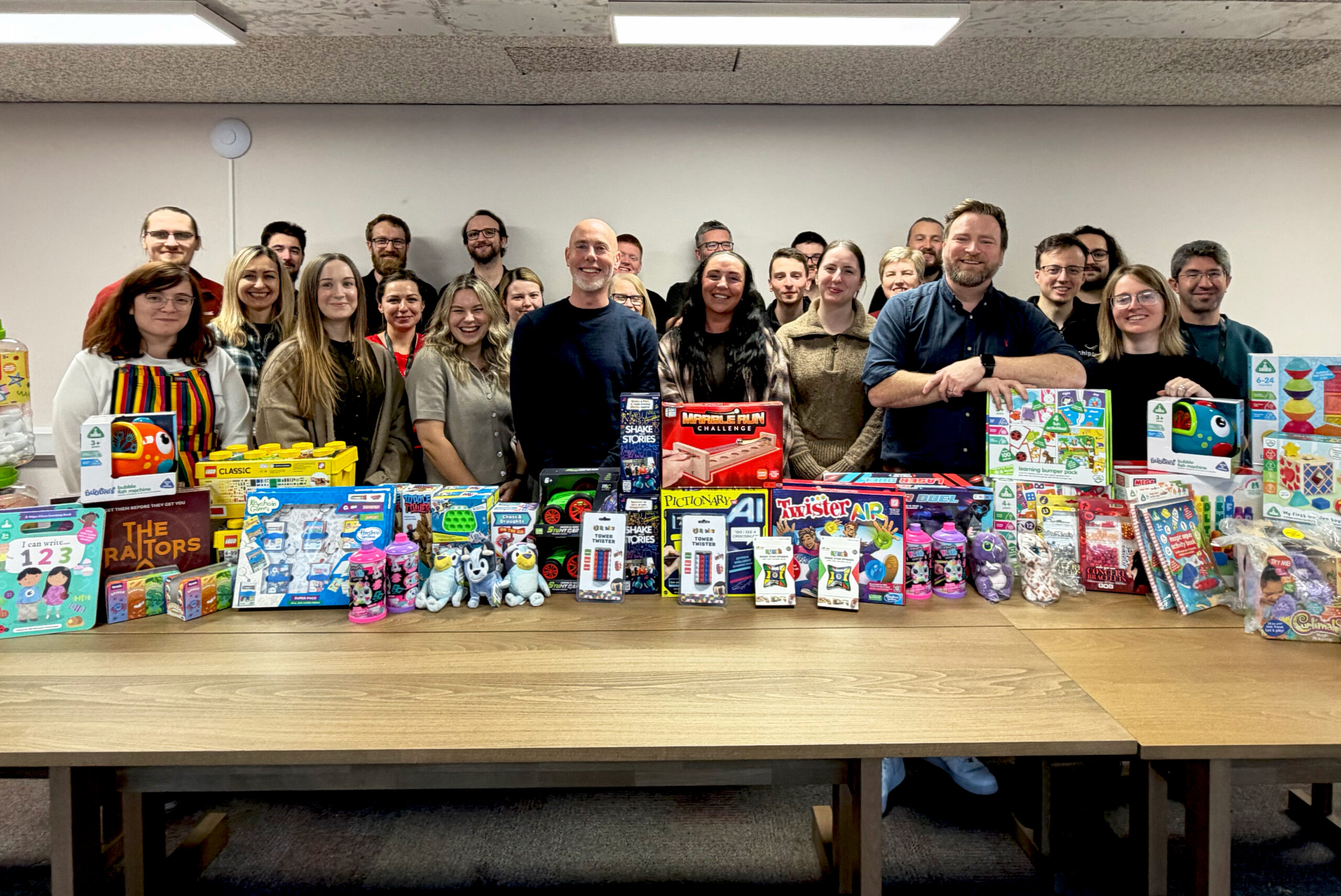A large group of people standing behind a long table which is stacked up with toys for children. Everyone is smiling at the camera.