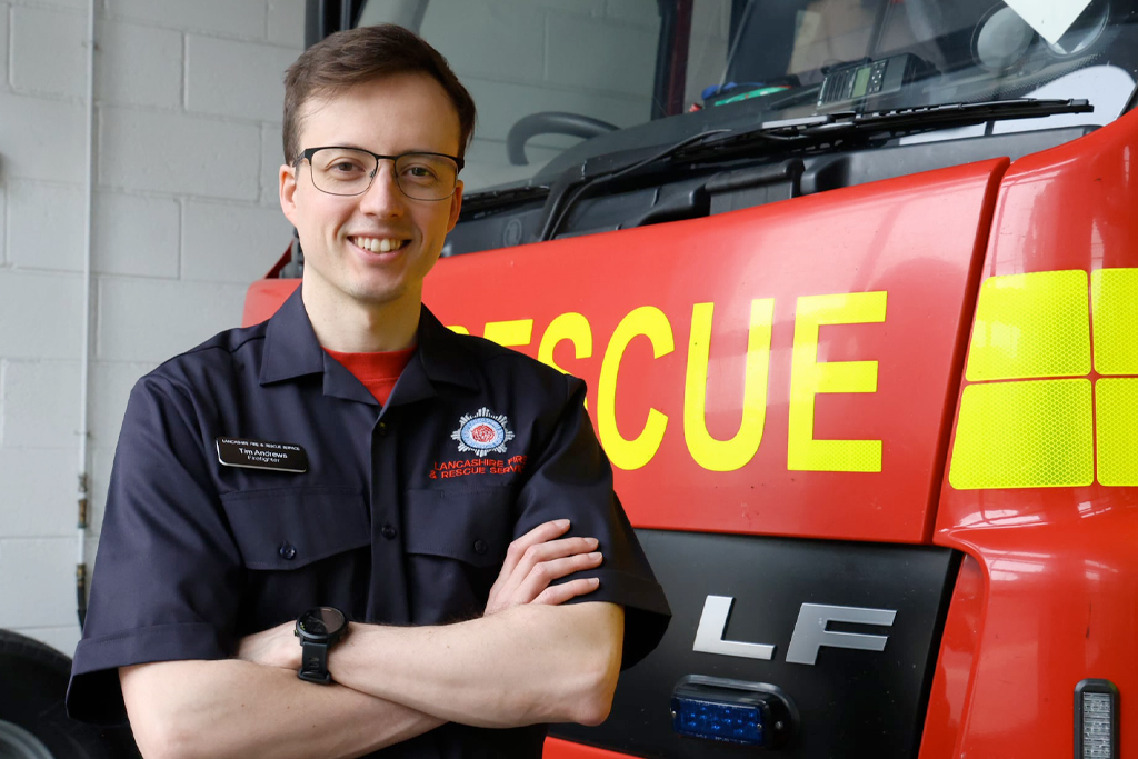 A man with short, dark hair and glasses stands on the left of the image in a blue duty-rig uniform displaying the Lancashire Fire and Rescue Service badge. His arms are folded across his chest and he is smiling at the camera. Behind him is the front of a red fire engine.