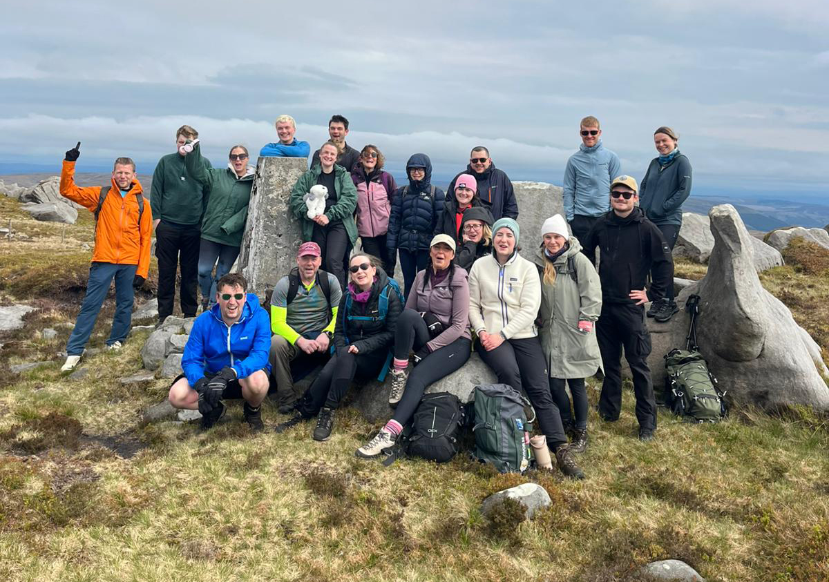 A group of people in outdoor clothing all stand together on a rocky outcrop on top of a hill. They are all smiling at the camera after completing a hike for charity.