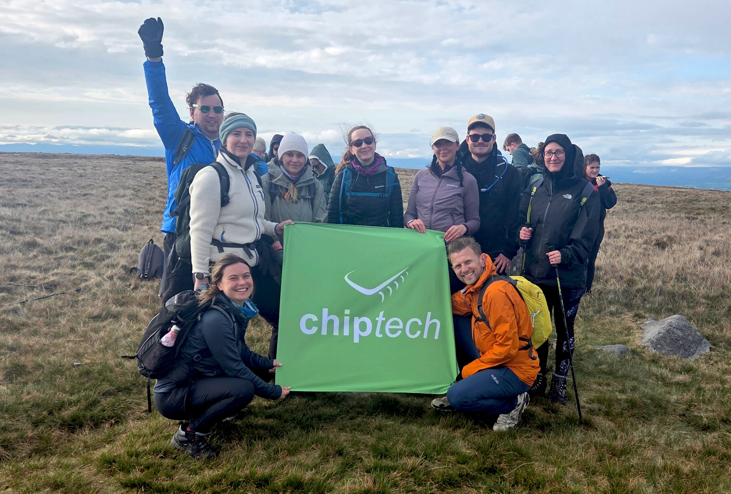 A group of people in outdoor hiking gear pose around a large green flag bearing a Chiptech logo after completing a 5-peak Challenge.