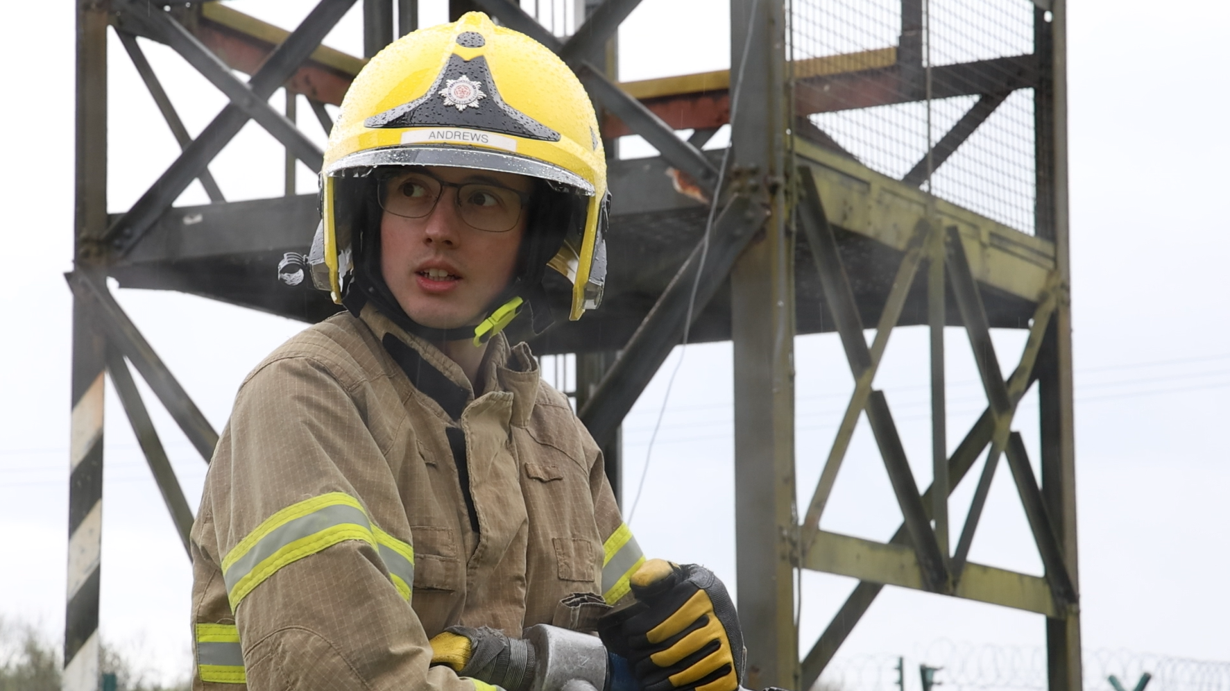 A firefighter stands outside holding a fire hose in his arms, with the branch in his hand. He is looking over his shoulder and wearing full PPE including a yellow helmet. Behind him is a metal drill tower.